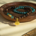 The Sandalwood & Green Jadeite Double-Strand Bracelet, featuring round beads and a pouch charm, is displayed on a wooden dish as sunlight casts gentle shadows across the elegant accessory.