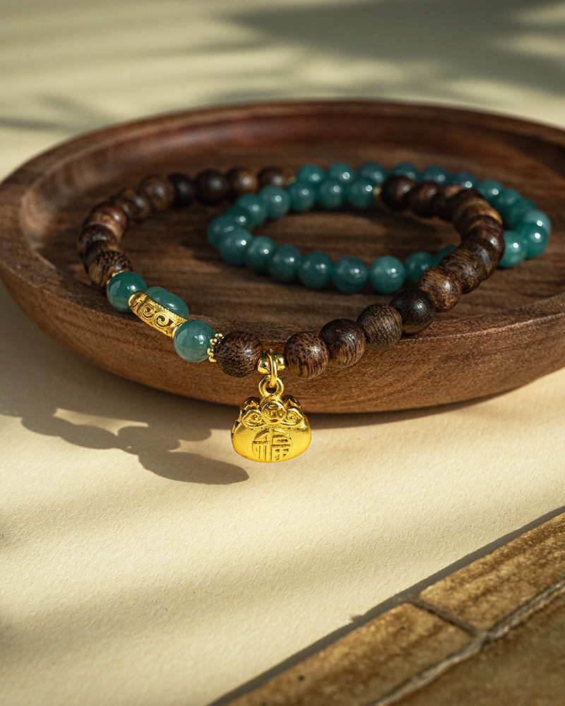 IMG_4757 The Sandalwood & Green Jadeite Double-Strand Bracelet, featuring round beads and a pouch charm, is displayed on a wooden dish as sunlight casts gentle shadows across the elegant accessory.