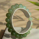 The Gradient Green Nephrite Jade Bracelet, featuring natural round beads in celadon-to-olive tones, is displayed upright on a white stand with gentle light and plant shadows in the background.