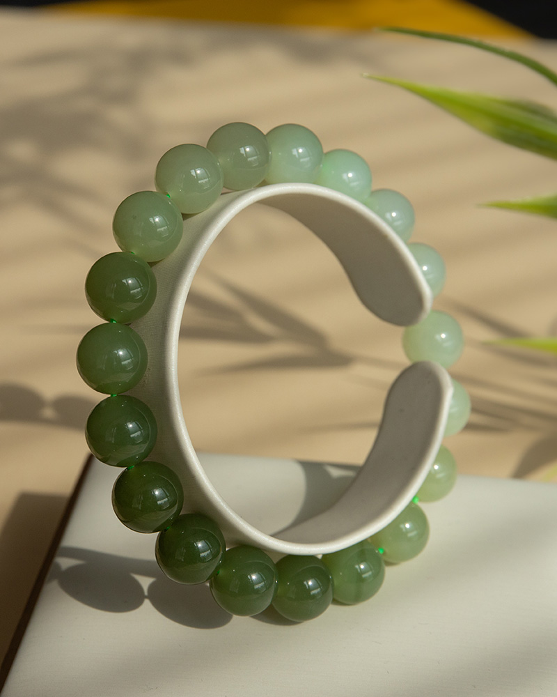 The Gradient Green Nephrite Jade Bracelet, featuring natural round beads in celadon-to-olive tones, is displayed upright on a white stand with gentle light and plant shadows in the background.
