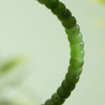 A close-up of the Natural Green Nephrite Jade Bracelet—handmade with polished, round, slightly translucent beads—set against a soft, blurred green background. Suitable stretch bracelet for men and women.