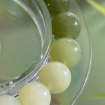 Close-up of the Gradient Green Nephrite Jade Bracelet, showcasing natural stone round beads in celadon-to-olive greens and off-white, resting on a clear circular surface with soft light and reflections.
