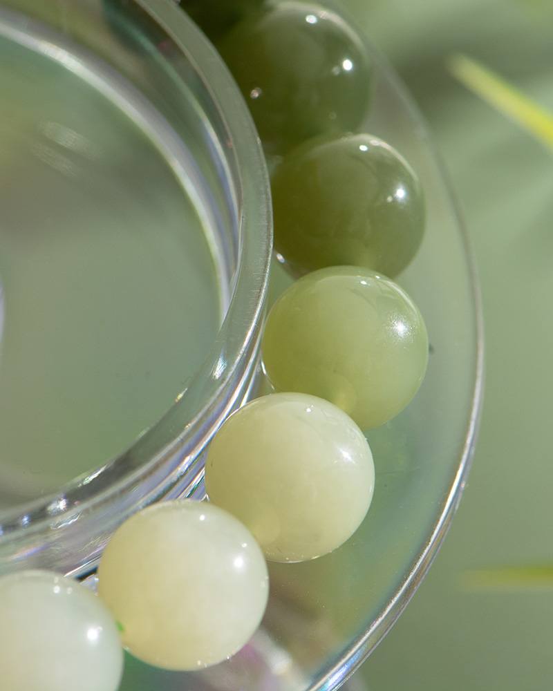 Close-up of the Gradient Green Nephrite Jade Bracelet, showcasing natural stone round beads in celadon-to-olive greens and off-white, resting on a clear circular surface with soft light and reflections.
