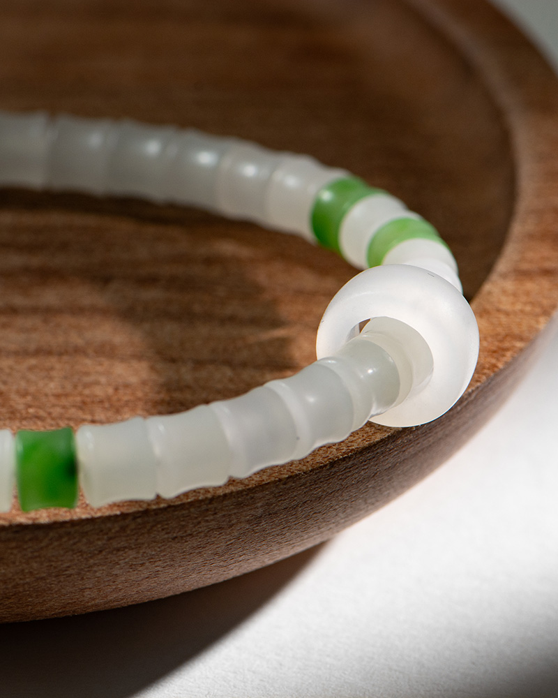 LYU_3896 A close-up of the Natural white Nephrite Jade Bracelet – Handmade Beads Stretch Bracelet with frosted white and green beads, displayed on a smooth wooden dish in gentle natural light that highlights its textured beauty.