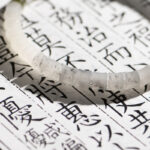 A White with Purple Veining Nephrite Jade Bracelet—featuring disc beads and Hetian Biyu accents—rests atop a page of vertical black Chinese characters, sunlight casting subtle shadows over the scene.