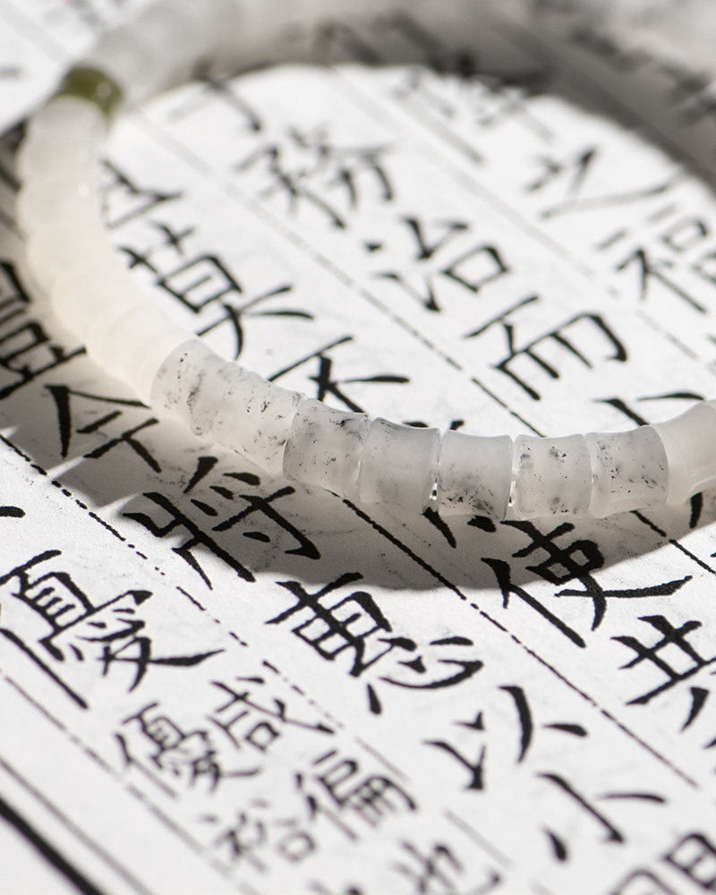 A White with Purple Veining Nephrite Jade Bracelet—featuring disc beads and Hetian Biyu accents—rests atop a page of vertical black Chinese characters, sunlight casting subtle shadows over the scene.