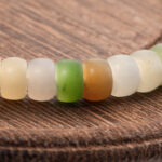 A close-up of the Pastel Spectrum Nephrite Jade Bracelet – Heishi Rondelles, featuring round, translucent beads in cream, green, and brown tones resting on a textured wooden surface.