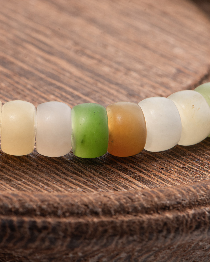 LYU_3930 A close-up of the Pastel Spectrum Nephrite Jade Bracelet – Heishi Rondelles, featuring round, translucent beads in cream, green, and brown tones resting on a textured wooden surface.