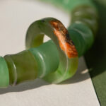 Close-up of the Nephrite Jade Bead Bracelet – Unique Ring with Natural Jade Skin, featuring green beads and one bead displaying an irregular brownish-red natural jade skin, shown on a white and green background.