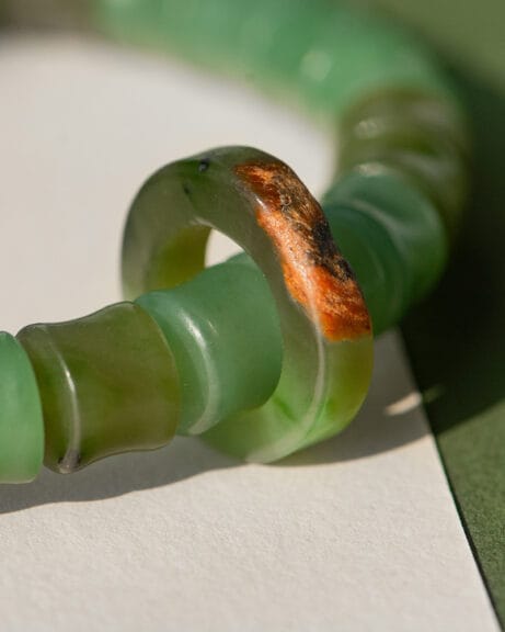 Close-up of the Nephrite Jade Bead Bracelet – Unique Ring with Natural Jade Skin, featuring green beads and one bead displaying an irregular brownish-red natural jade skin, shown on a white and green background.