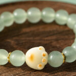 The Mint Green Aventurine Quartz Bracelet features pale green round beads and a cream cat paw charm with a yellowish spot, displayed on a wooden surface.