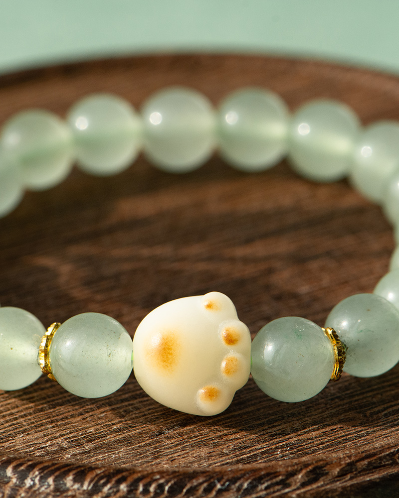 LYU_3948 The Mint Green Aventurine Quartz Bracelet features pale green round beads and a cream cat paw charm with a yellowish spot, displayed on a wooden surface.