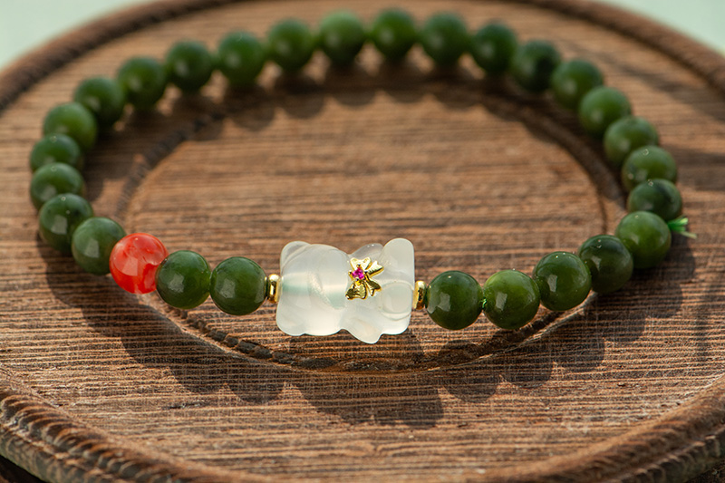 LYU_3956 The Deep Green Nephrite Jade Bracelet features round beads, a red agate accent, and a Yanyuan bear charm with gold and pink flower detail, shown on a wooden surface.