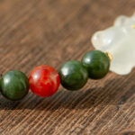 A close-up of the Deep Green Nephrite Jade Bracelet, featuring round beads, a translucent red agate accent, and a white Yanyuan bear charm, displayed on textured wood.