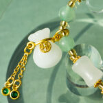 Close-up of the Icy Green Tianshan Cui Chalcedony Bracelet featuring bamboo-knot beads, a white lucky pouch charm, a gold coin with Chinese characters, and two green gemstone dangles displayed on glass.