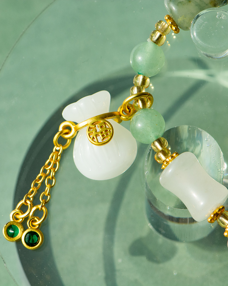 LYU_3968 Close-up of the Icy Green Tianshan Cui Chalcedony Bracelet featuring bamboo-knot beads, a white lucky pouch charm, a gold coin with Chinese characters, and two green gemstone dangles displayed on glass.