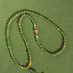 The Olive Green Nephrite Jade Wrap Bracelet – Round Beads, Convertible Necklace features green beads with a pink accent and gold details, coiled on a textured surface. A larger bead and tassel adorn one end.