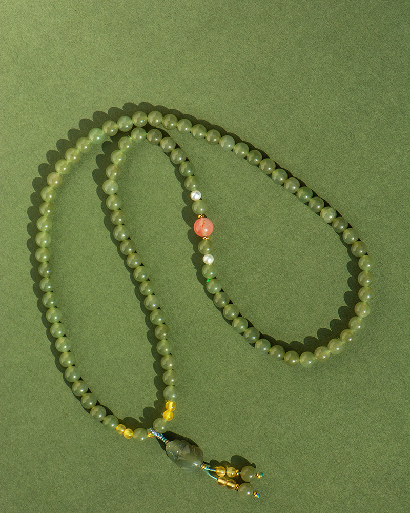 LYU_3980 The Olive Green Nephrite Jade Wrap Bracelet – Round Beads, Convertible Necklace features green beads with a pink accent and gold details, coiled on a textured surface. A larger bead and tassel adorn one end.