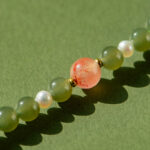 A close-up of the Olive Green Nephrite Jade Wrap Bracelet, featuring round green jade beads and small white pearls, with a larger peach bead with red speckles, displayed against a green background.