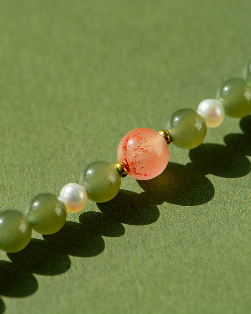 LYU_3995 A close-up of the Olive Green Nephrite Jade Wrap Bracelet, featuring round green jade beads and small white pearls, with a larger peach bead with red speckles, displayed against a green background.
