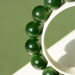 Close-up of the Deep Green Nephrite Jade Bracelet with 12mm polished round beads, photographed on a light and green background, sunlight casting soft shadows.