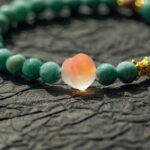 The Green Jadeite Bracelet features round green beads with a clear Buddha-head charm at the center, elegantly displayed on a textured dark surface.