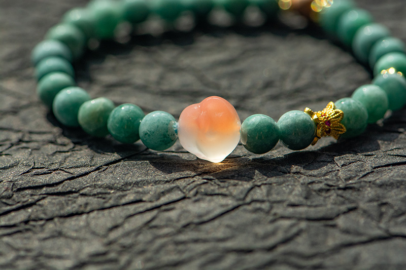 LYU_4007 The Green Jadeite Bracelet features round green beads with a clear Buddha-head charm at the center, elegantly displayed on a textured dark surface.