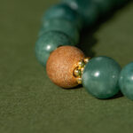 Close-up of the Green Jadeite Bracelet featuring round green beads, a clear Buddha-head charm, and gold spacers, displayed on a textured green surface.
