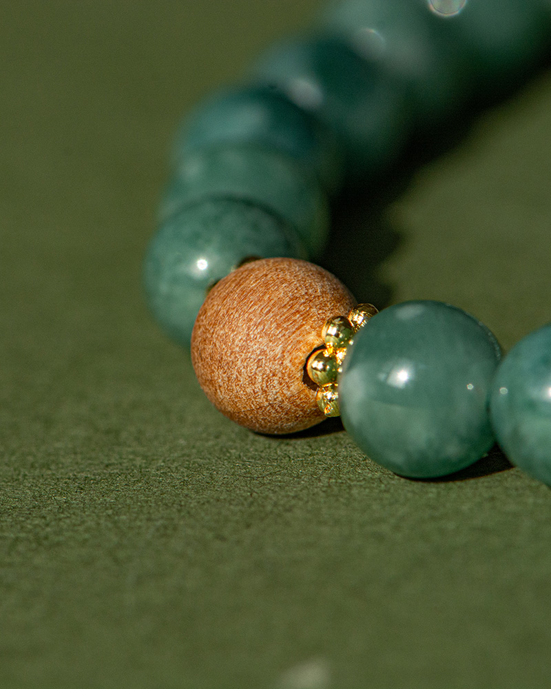 LYU_4011 Close-up of the Green Jadeite Bracelet featuring round green beads, a clear Buddha-head charm, and gold spacers, displayed on a textured green surface.