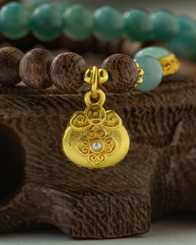 LYU_4012 A close-up of the Sandalwood & Green Jadeite Double-Strand Bracelet, featuring round green jadeite and sandalwood beads with a gold-tone pouch charm, displayed on a textured wooden surface.
