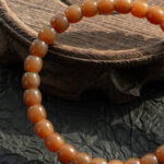A close-up of the Natural Hetian Jade Beaded Bracelet, featuring round brownish-orange beads for meditation and energy, resting on a textured wooden surface with part of the bracelet on a dark, crinkled background.