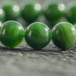 Close-up of a Deep Green Nephrite Jade Bracelet with 12mm polished round beads, displayed on a textured gray surface. Soft lighting and shallow depth of field highlight the rich color and shine of the stones.