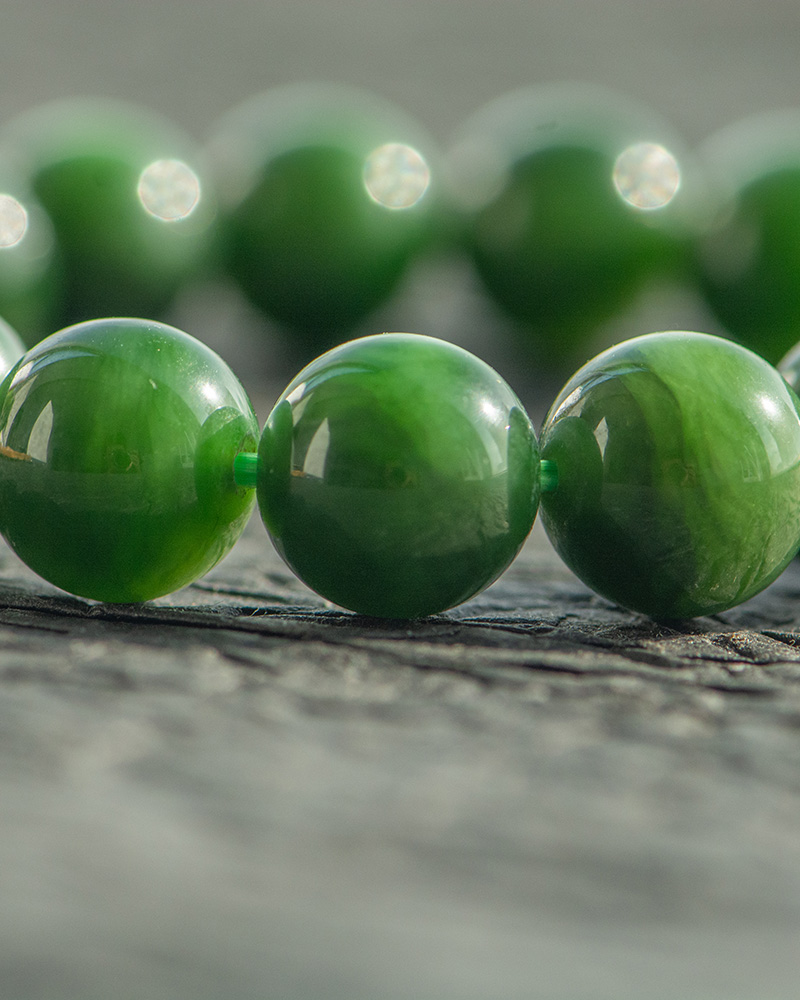 Close-up of a Deep Green Nephrite Jade Bracelet with 12mm polished round beads, displayed on a textured gray surface. Soft lighting and shallow depth of field highlight the rich color and shine of the stones.