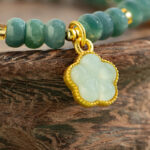 A close-up of the Teal Green Jadeite Bracelet, featuring disc beads and trio charms, displayed on a carved wooden surface.