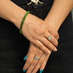 Close-up of hands with teal nails and silver rings, wearing a Natural Green Nephrite Jade Bracelet – Handmade Polished Round Beads Stretch Bracelet for Men and Women, paired with black pants and a belt with a large heart-shaped buckle.