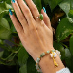 A hand with blue-painted nails wears a green stone ring and the Icy Green Tianshan Cui Chalcedony Bracelet—featuring bamboo-knot beads and a white lucky pouch charm—resting on green leafy plants.