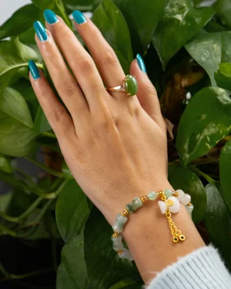 A hand with blue-painted nails wears a green stone ring and the White Quartzite Jade Bracelet – featuring carved pillow beads and a gold-tone tassel charm – resting on green leafy plants.