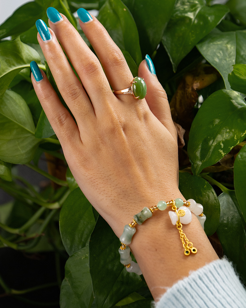 LYU_4175 A hand with blue-painted nails wears a green stone ring and the Icy Green Tianshan Cui Chalcedony Bracelet—featuring bamboo-knot beads and a white lucky pouch charm—resting on green leafy plants.
