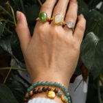 A hand wearing three gemstone rings and the Sandalwood & Green Jadeite Double-Strand Bracelet with a pouch charm rests on green leafy plants. The person is in a light blue sleeve.