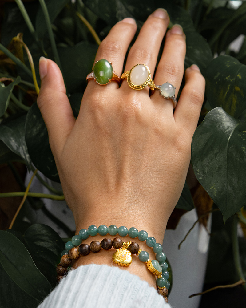 LYU_4194 A hand wearing three gemstone rings and the Sandalwood & Green Jadeite Double-Strand Bracelet with a pouch charm rests on green leafy plants. The person is in a light blue sleeve.