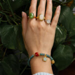 A hand adorned with three gemstone rings and the Teal Green Jadeite Bracelet – Disc Beads with Trio Charms is posed before leafy green plants. The wearer is dressed in a light blue ribbed sweater.