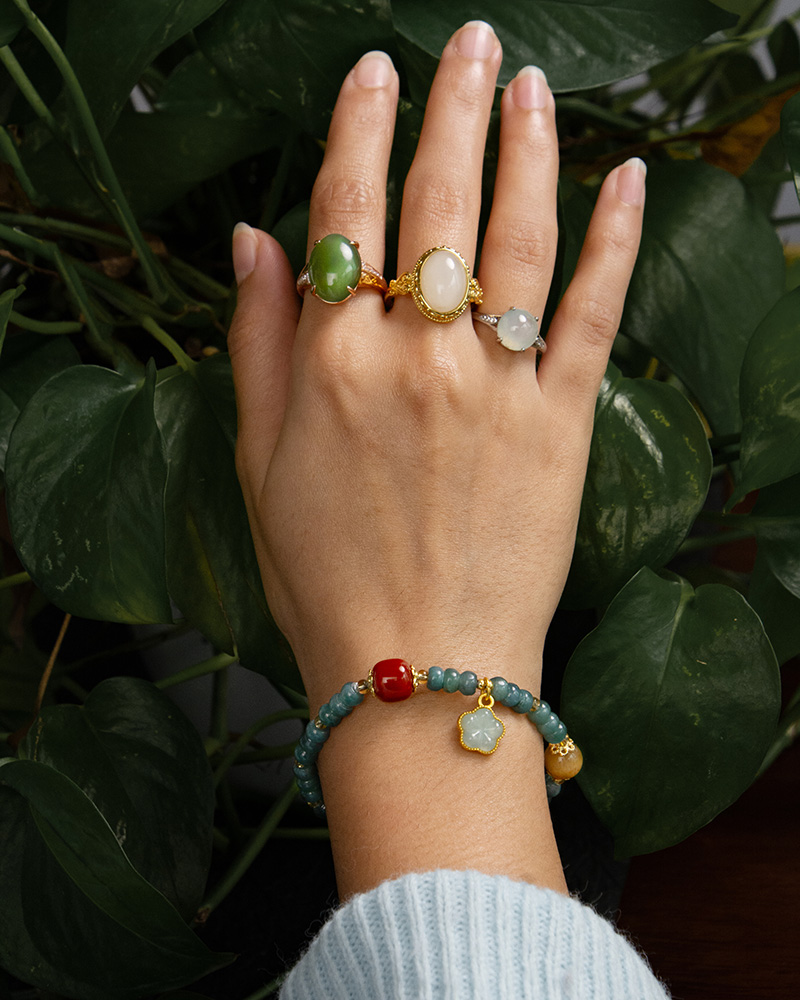 LYU_4198 A hand adorned with three gemstone rings and the Teal Green Jadeite Bracelet – Disc Beads with Trio Charms is posed before leafy green plants. The wearer is dressed in a light blue ribbed sweater.