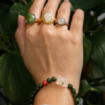 A hand adorned with three gemstone rings and the Deep Green Nephrite Jade Bracelet—featuring round beads, a red agate accent, and a Yanyuan bear charm—is posed in front of lush green plants.