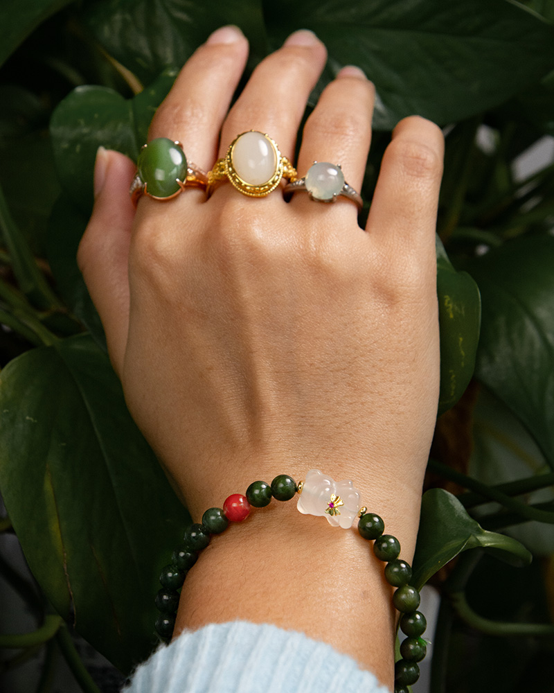 LYU_4199 A hand adorned with three gemstone rings and the Deep Green Nephrite Jade Bracelet—featuring round beads, a red agate accent, and a Yanyuan bear charm—is posed in front of lush green plants.
