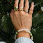 A hand with three stone rings, including the Hetian White Nephrite Jade Ring – Oval Cabochon in a gold-tone filigree setting, and a white-green beaded bracelet, is held over green leaves. The person wears a light blue sleeve.