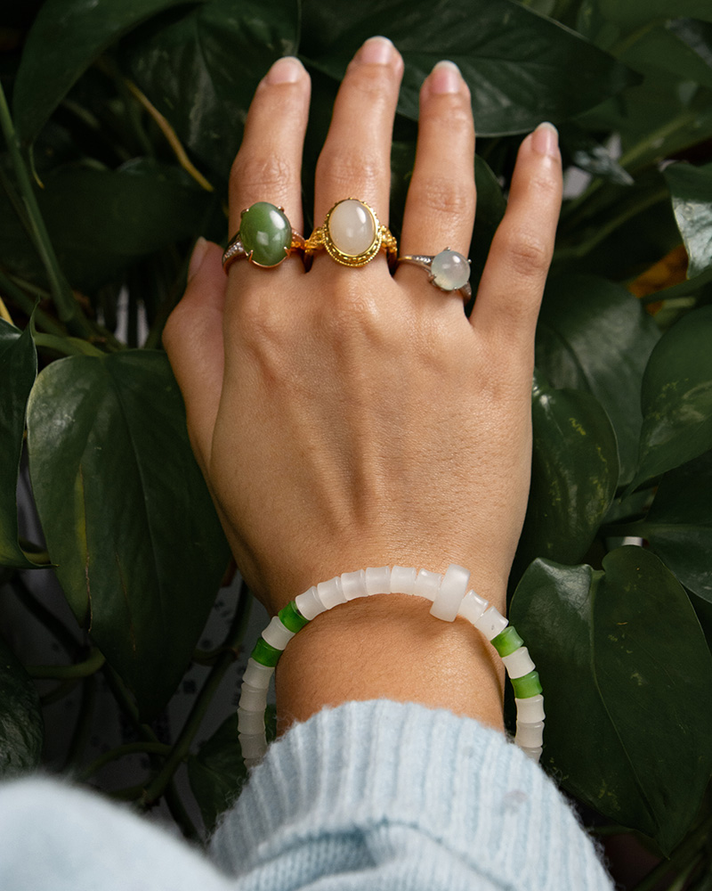A hand with three stone rings, including the Hetian White Nephrite Jade Ring – Oval Cabochon in a gold-tone filigree setting, and a white-green beaded bracelet, is held over green leaves. The person wears a light blue sleeve.