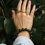 A hand adorned with three rings and the Deep Green Nephrite Jade Bracelet – 12mm Polished Round Beads is raised before leafy green plants. The person wears a light blue sweater.