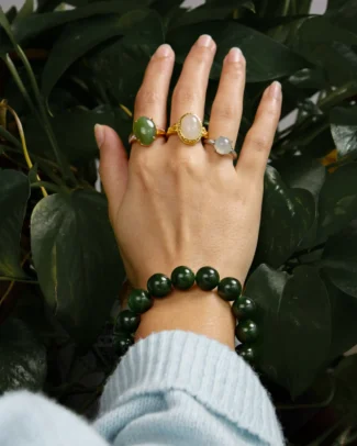 A hand adorned with three gemstone rings and the Deep Green Nephrite Jade Bracelet – Spherical Beads, High-Gloss Polish is shown against a backdrop of green leaves, with the wearer dressed in a light blue sweater.