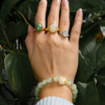 A hand in a light blue, ribbed sleeve wears three gemstone rings and the Mint Green Aventurine Quartz Bracelet with round beads and a cat paw charm, held over lush green leaves. The jewelry highlights green, white, and gold tones.