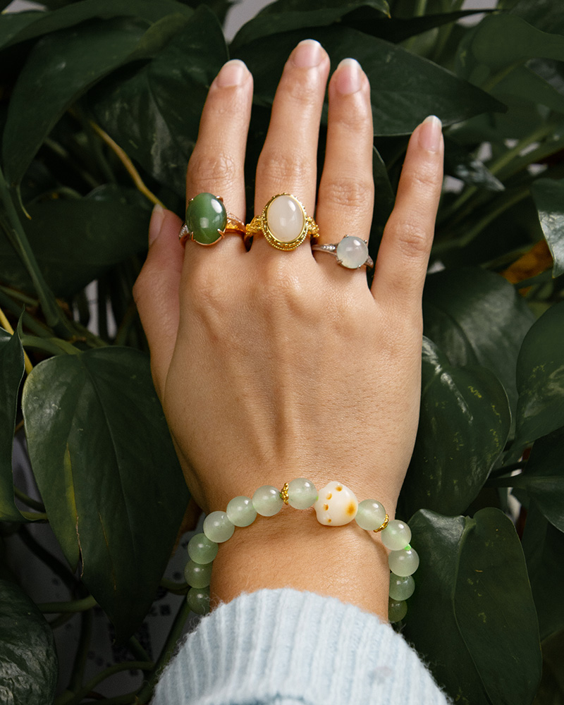 LYU_4213 A hand in a light blue, ribbed sleeve wears three gemstone rings and the Mint Green Aventurine Quartz Bracelet with round beads and a cat paw charm, held over lush green leaves. The jewelry highlights green, white, and gold tones.